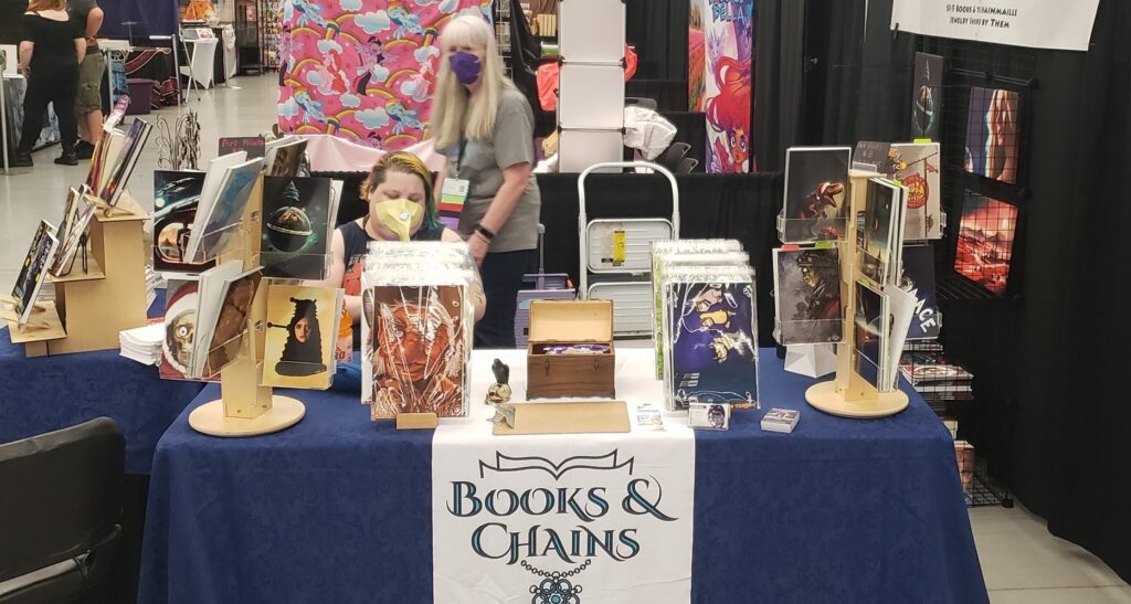 Photo of the Books & Chains booth at GeekGirlCon 2023. This is a site shot showing off some of Raven Oak's artwork.