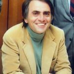 Carl Sagan, wearing a mustard-colored jacket and a mint green turtleneck. He is smiling and has dark brown hair with a little bit of gray.