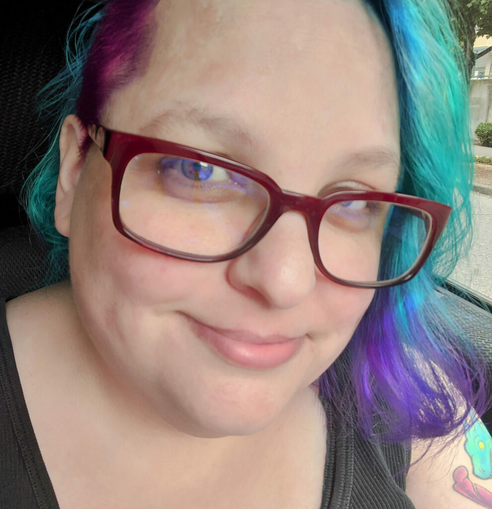 Author & artist Raven Oak, pictured with teal & purple hair, a side shave, and red glasses. They are smiling.